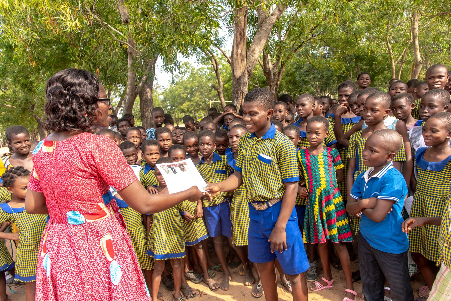 Lorlor, the social worker from Dove child development center, in a pink dress, is handing out letters. The children are gathered around wearing colorful green and blue outfits.
