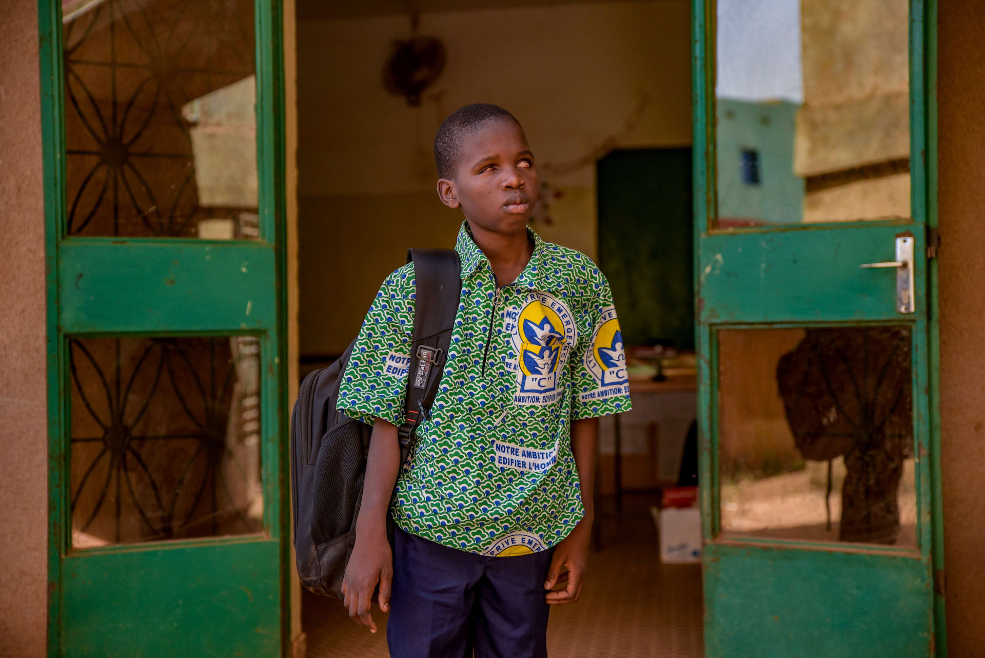 Kader is wearing a blue, white, and green patterned shirt and is holding a black back pack. He is standing in front of his classroom. The building is brown and the doors are green.