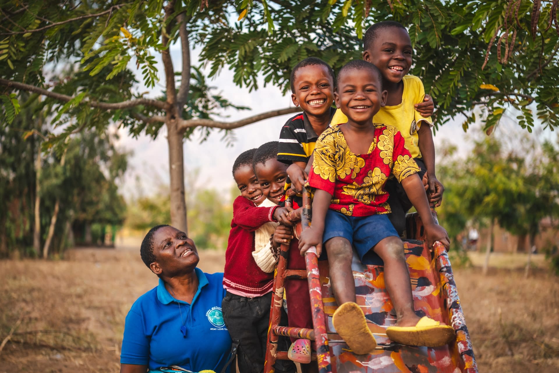 A group of 5 children crowd onto a playground slide, smiling together. Pastor Beauty Senjele stands next to the slide, looking up at the children.