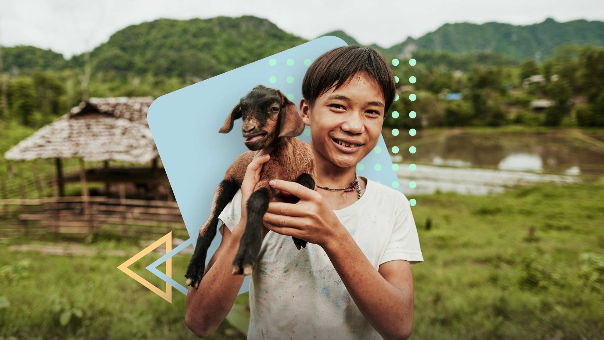 An Asian boy stands in a green field holding a brown goat on his shoulder.