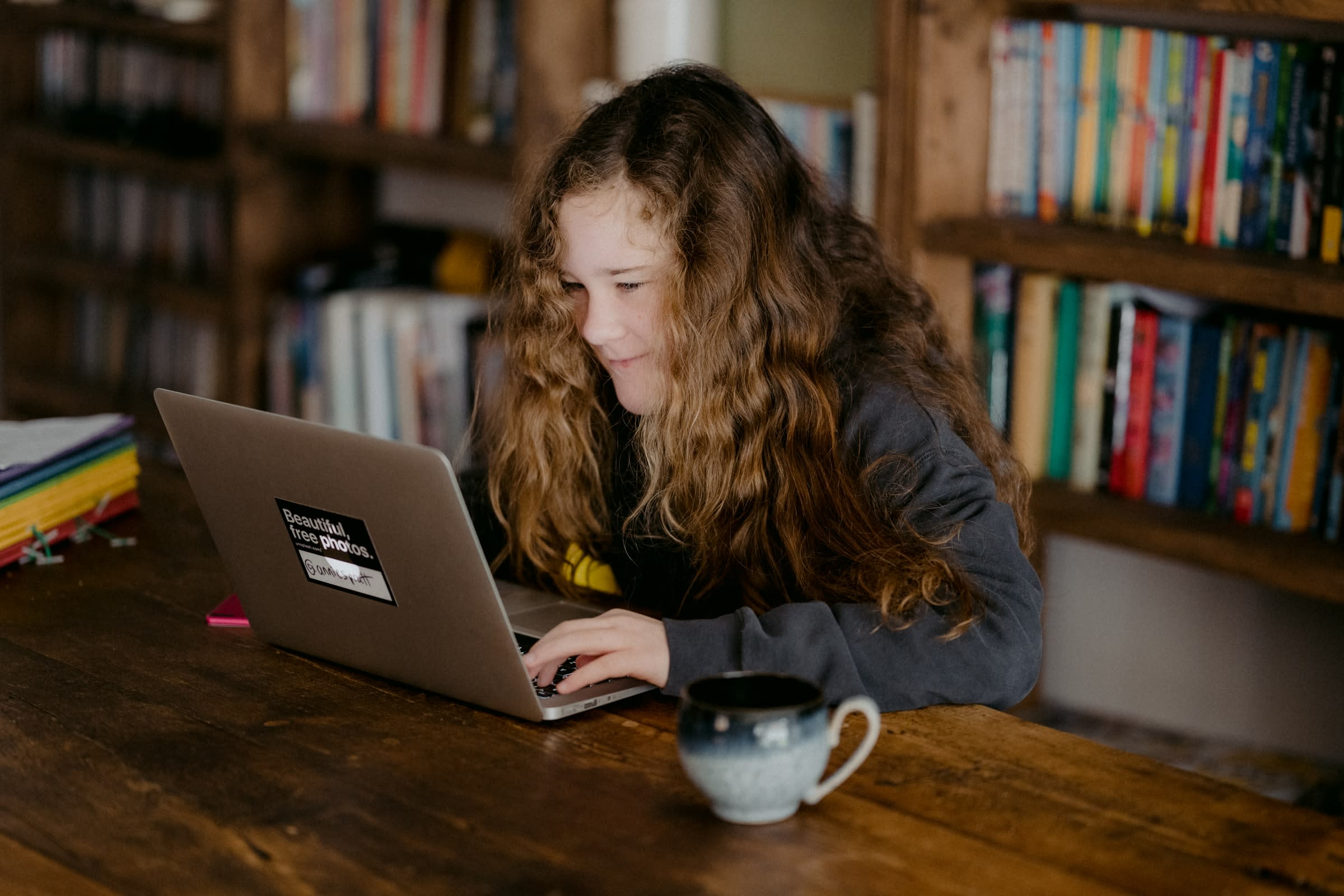 A girl with curly hair smiles at her laptop in front of a bookcase