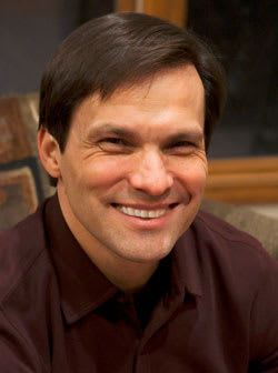 A man in a brown shirt and dark hair smiles at the camera.