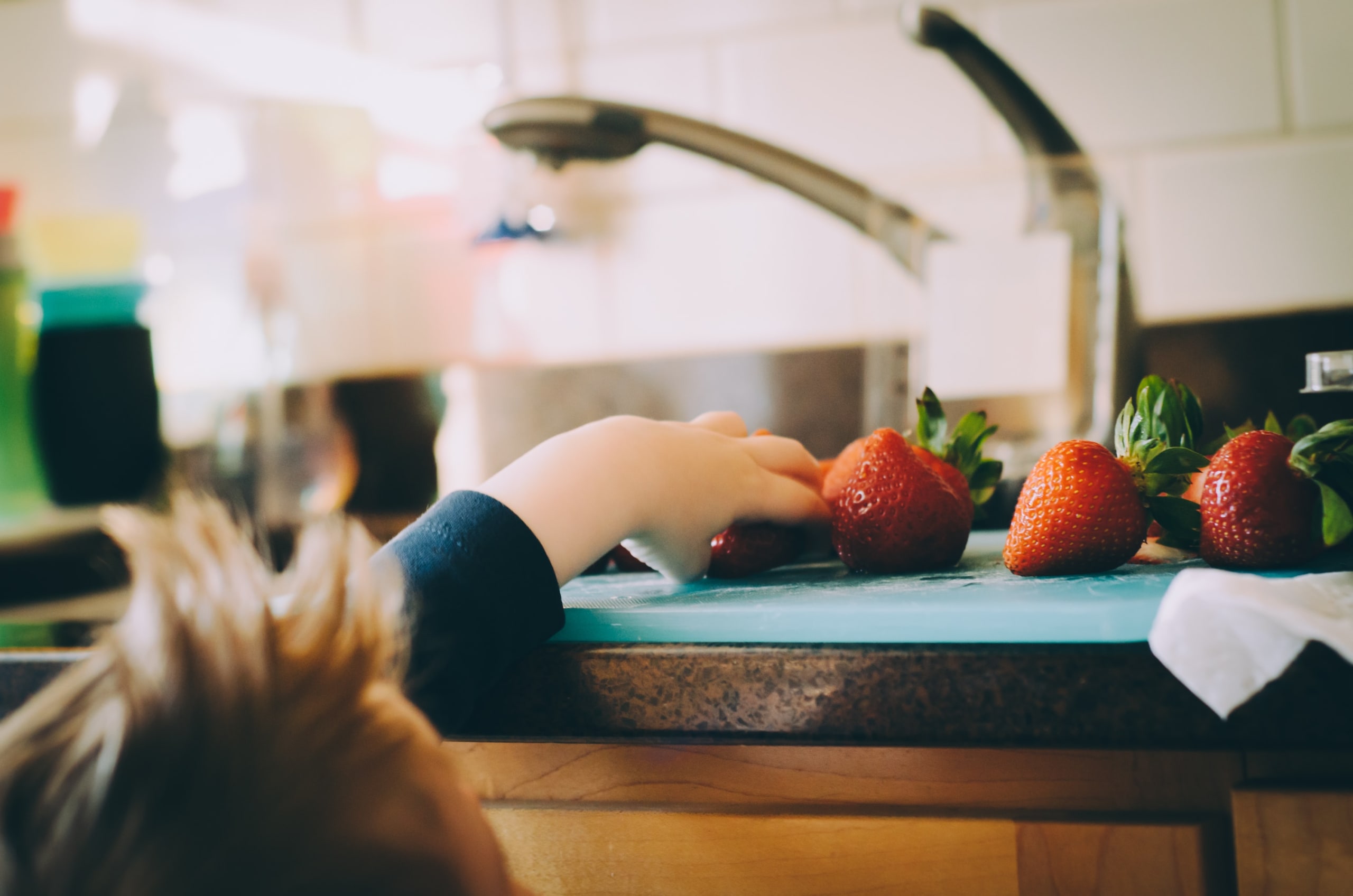 A little boy reaches up to a counter to reach for strawberries. There is a sink beside him.