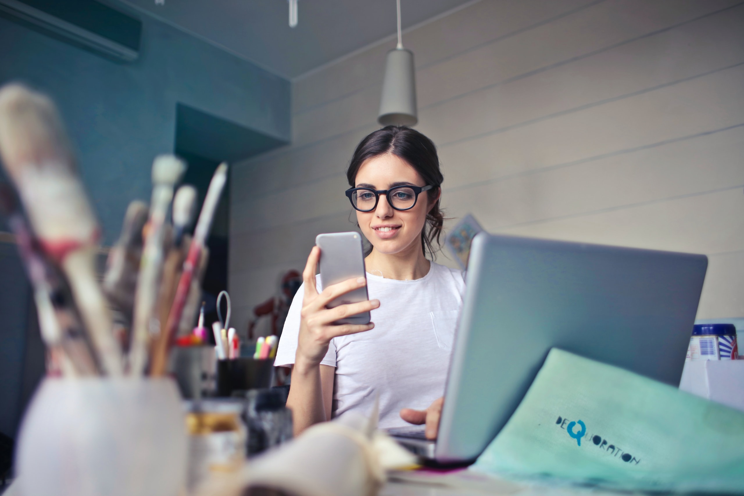 Woman sits in front of a computer and holds up her cellphone