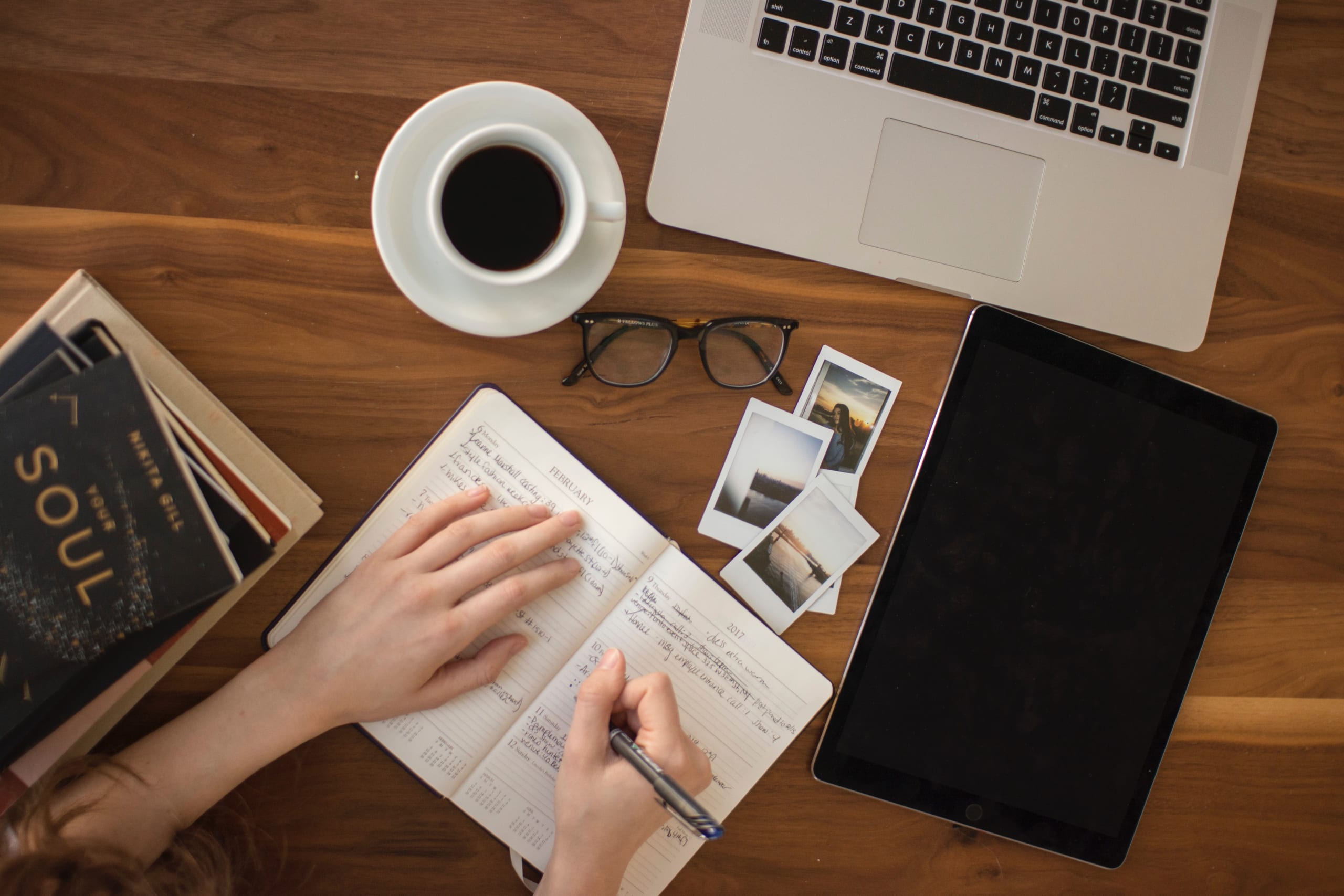 Hands writing in a notebook on a desk, surrounded by a laptop, reading glasses, books and a cup of coffee.