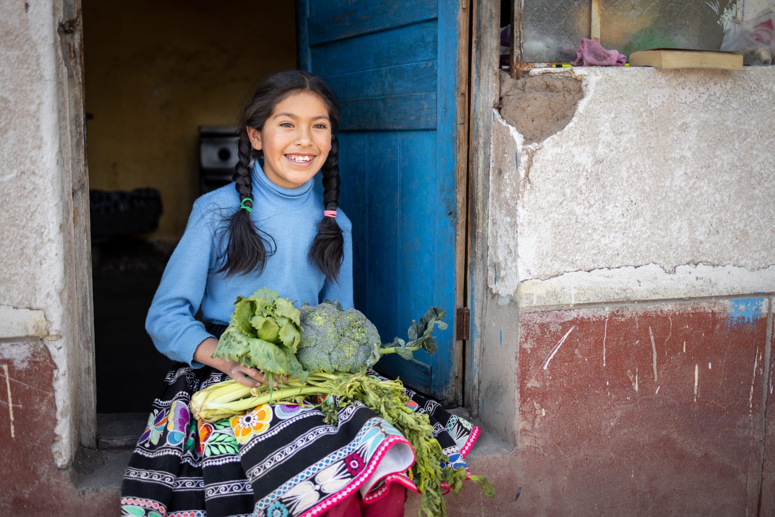 A young girl in a blue top and colourful skirt sits in the doorway of her home, holding the vegetables she has grown on her lap
