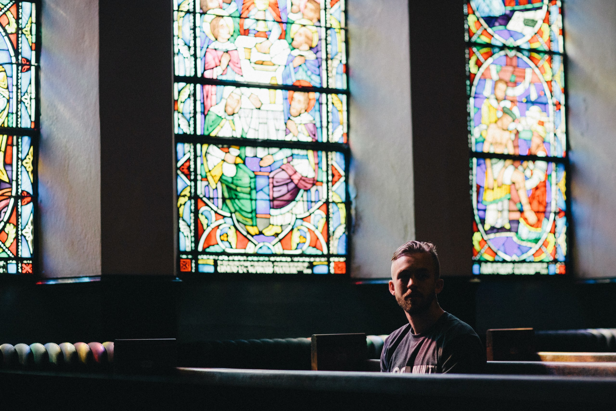 a young man sits in a church pew pondering, with a background of stained glass windows