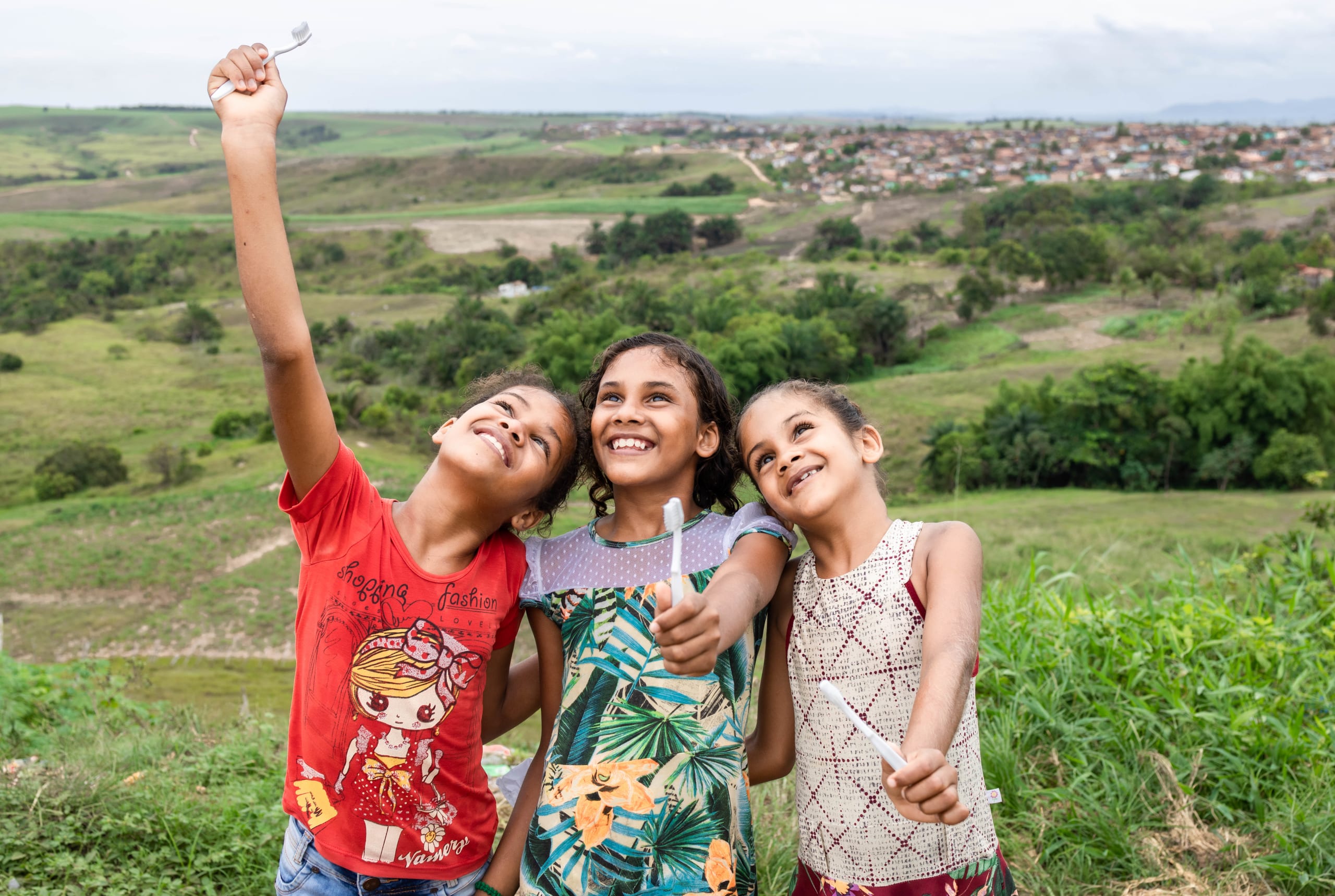 Three girls standing on a hill hold up toothbrushes and smile.