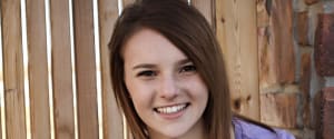 A young girl with brown hair smiles in front of a wooden fence.