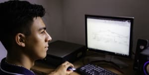A teenager sits infront of a computer, his hand on the mouse. He looks at the screen.