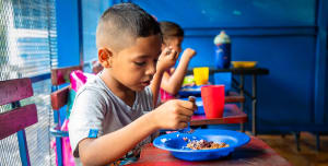 Grade school aged kids sit at a table and eat food from blue bowls.