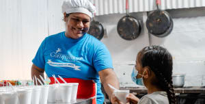 A lady wearing a blue shirt and a chef hat smiling at a little girl
