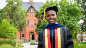 A man stands in a graduation gown and cap. He is smiling and standing in front of a university.