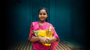A South Asian girl holds groceries in her arms. She is wearing a red dress and pink scarf.