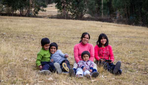 A family from Peru sits on a grass hill smiling at the camera. There are two boys, 2 girls and a mom