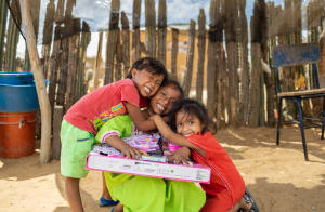 In Colombia, Eldimar is wearing a bright green dress. She is sitting down outside her home and is unwrapping her gifts from her sponsor. Her sisters, both wearing red, are hugging her.