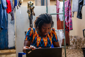 Winner, a young woman in Togo, is wearing an orange and blue dress. She is inside her home and is working on her computer.