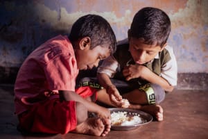 Two boys sit on the floor and share a bowl of food.