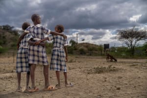 three school girls in their uniforms standing together, arms around each other, as they stare off into the horizon