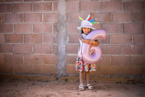 A girl in a colourful dress holds a balloon six and wears a birthday hat