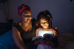 A young dark skinned child looks at her Bible, sitting beside her mother. The room is dark, but the Bible is glowing with light.