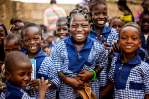 A group of children from Burkina Faso smile and laugh. They are wearing blue and white school uniforms and are standing in front of a Compassion centre building