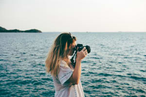 A woman stands by the water and takes a photograph.