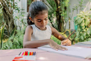 Little girl sits at a desk with a pink table cloth. She has coloured pencils beside her and is writing a letter.