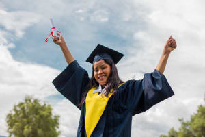 A young woman with a graduation cap and dark gown celebrates and holds her diploma up in the air