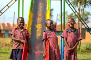 Three triplets smile together by a colourful slide. They are wearing matching red outfits.