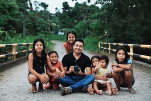 A man sits cross-legged on a road with a camera in his hands.He is surrounded by smiling kids.
