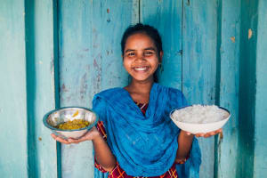 A young woman stands in front of a blue background holding two bowls of rice and beans. She is wearing a blue scarf and smiling.