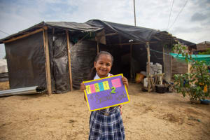 Sara stands in front of her house wearing a school uniform and holding a sign that says "First Day of Second Grade".