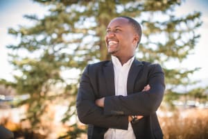 A man stands with his arms crossed as he looks out of frame. He smiles, seeming happy and proud.