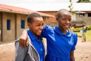 Two young girls, teens, teenagers stand outside the project with their arms around each other, hugging as they smile for the camera. They are both wearing the blue dress project uniform.