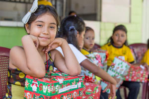 A close-up picture of a girl holding a Christmas gift. She is smiling and wearing a blue hair bow.