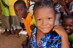 A girl in a blue shirt looks into the camera and smiles with bright eyes. A boy peers over her shoulder.
