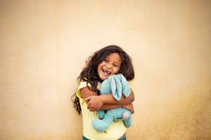 A little girl with long brown wavy hair smiles with missing teeth, snuggling a blue bunny. She is infront of a yellow wall.