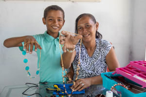 Boy in a turquiouse shirt and woman in a blue and white shirt pose at a table showing handmade jewelry to the camera.