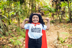 Valery is wearing a superhero cape and a t-shirt that says "Leukemia Warrior" and is holding her arms in a powerful pose. She is outside surrounded by trees.