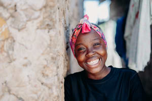 Portrait of Alice by a stone wall. She is wearing a black shirt and colorful pink scarf on her head. She is smiling at the camera.