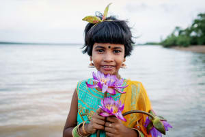 A young girl stands on the shore of the ocean in a colourful dress. She's holding a pink flower.