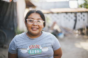 A girl wearing pink glasses and a grey t-shirt smiles.