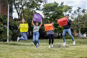 Hellen, Jhoselyn, Natalia and Samuel are outside jumping into the air over the grass while they hold motivational posters that say, "God loves you," "Serve your neighbor," and Love your neighbor."