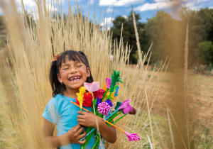 A young girl in a blue tshirt laughs while holding a bouquet of paper flowers.
