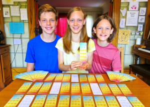 Three children stand together in front of a table. The middle one is holding a bookmark.