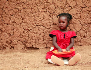 A little girl in a red dress sits against a mud wall and holds an empty plate.