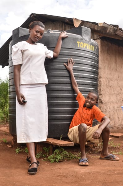 Thirteen-year-old Eric Njuki, a young teen boy, wearing an orange shirt and khaki shorts, at home, with his mother, Rose Nduta, adult female wearing a white shirt and skirt, are standing with a Compassion-provided water tank to harvest rain water, large black water reservoir, container. A walled mud home and greenery is in the background.