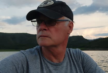 A man in a ball-cap and glasses looks over his shoulder in front of a sunset on water.