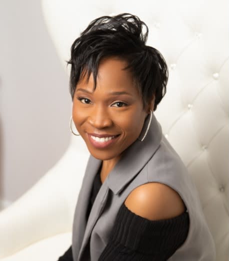 Compassion Ambassador, Donnett Thompson-Hall is sitting on a white chair and smiling. She is wearing a black shirt with a grey vest and hoop earrings.